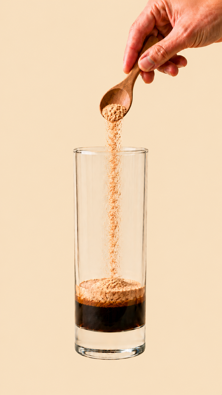 Glass with coffee, cinnamon and a fine layer of pale tan maca powder settled on top; a hand visible above holding a wooden teaspoon, releasing a fine stream of maca powder falling into the glass