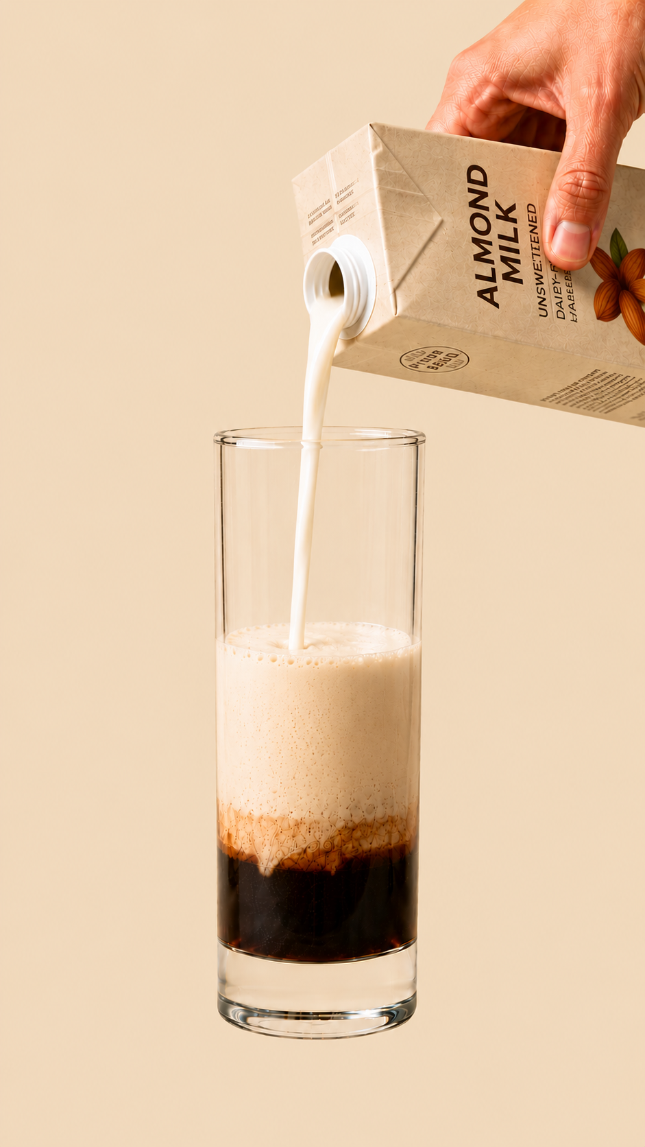 Glass with layered coffee, cinnamon, maca and warm almond milk filling the upper portion; a hand visible above holding a small glass jug, pouring warm almond milk into the glass