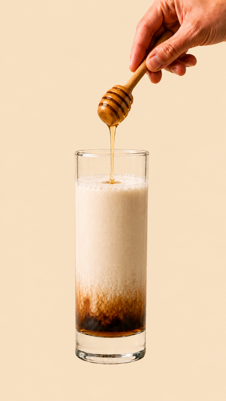 Glass with full latte layers and a thin amber ribbon of raw honey settled across the top; a hand visible above holding a wooden honey dipper, drizzling raw honey into the glass